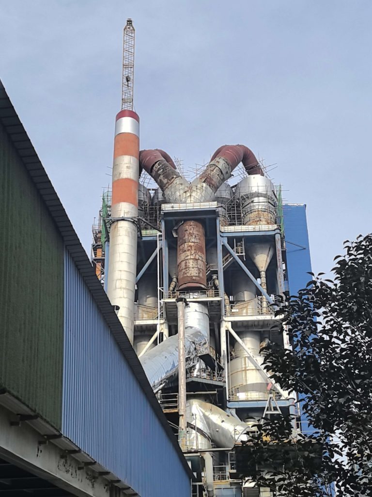 c1 cyclone retrofit bbmg wenxi cement 5000ton per day - Nanjing Sequoia Industrial Solutions Co.,ltd Industrial refinery structure with tall smokestack and a network of curved metal pipes against a clear blue sky, adjacent to a blue building edge.